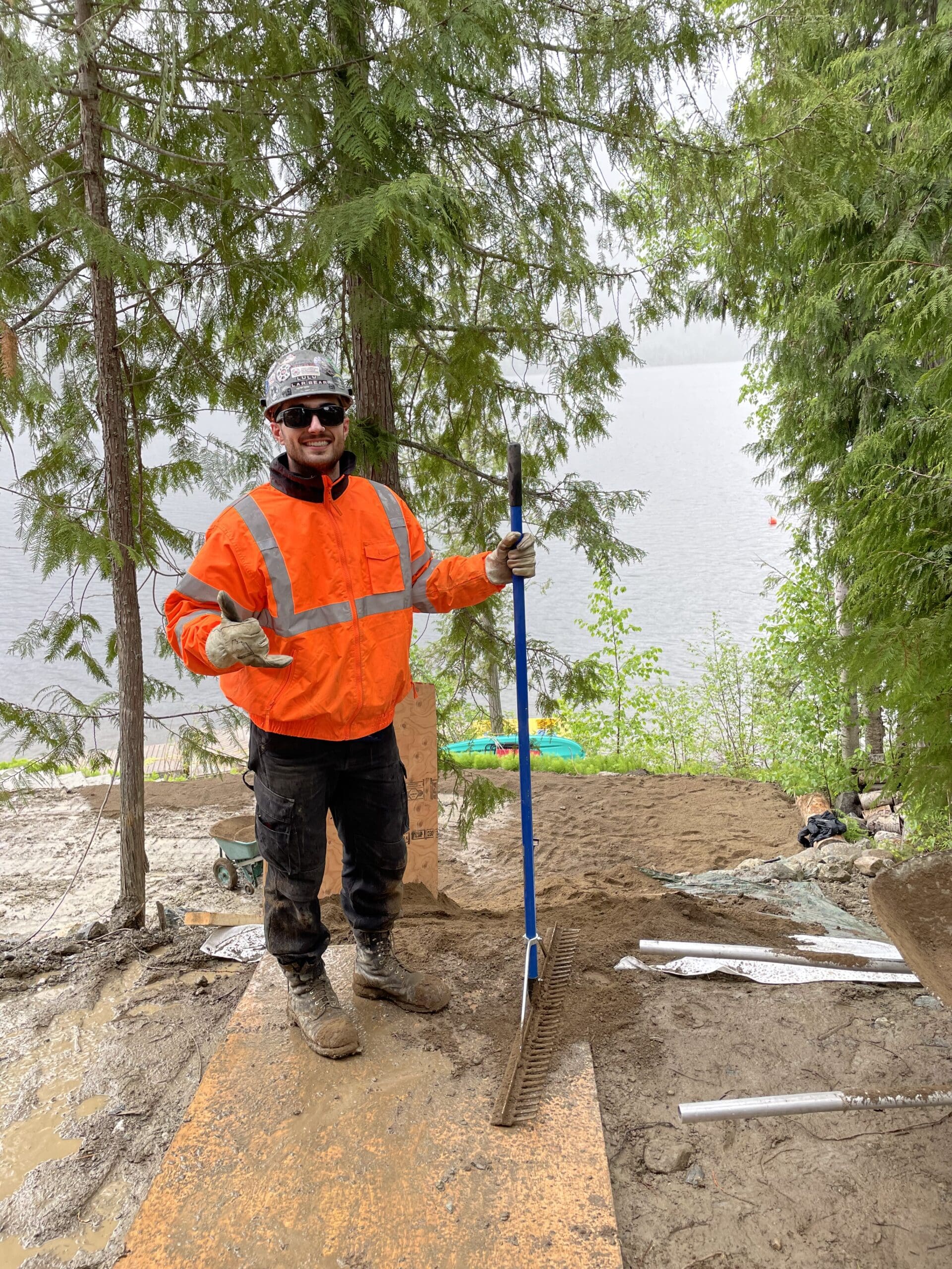 By the Yard employee in full protective gear holding a rake in front of a lake