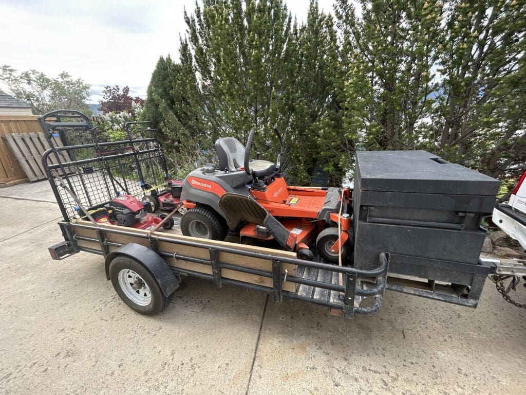 A utility trailer loaded with professional lawn care equipment, including a Husqvarna zero-turn mower, a red push mower, and a large black storage box. The trailer is parked on a concrete driveway next to a house and surrounded by trees and shrubs.