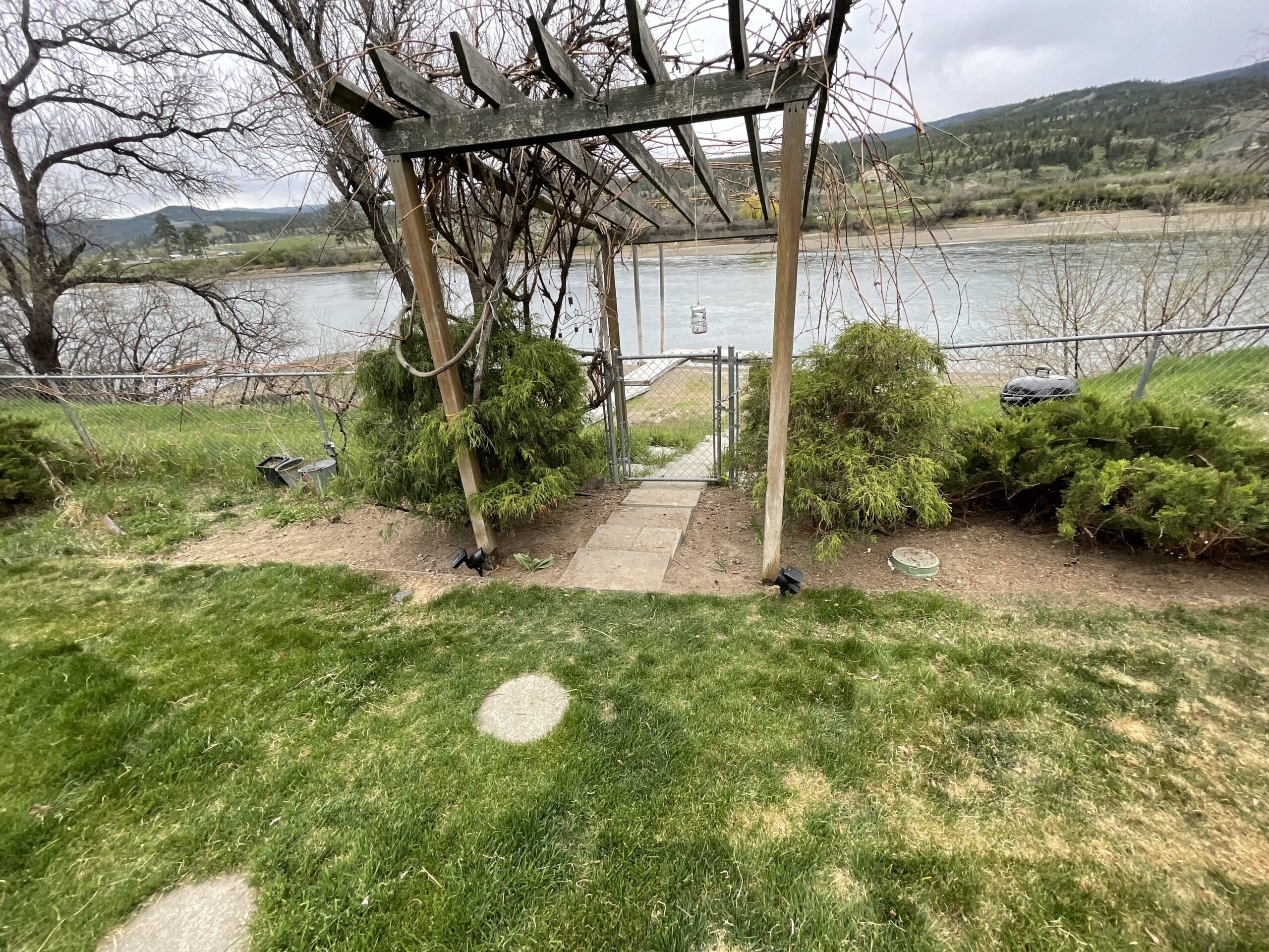 A weathered wooden pergola stands over a stone path leading to a gated fence along a riverbank. Surrounded by sparse lawn, evergreen shrubs, and bare trees, the scene overlooks a wide, calm river with forested hills in the distance under an overcast sky.