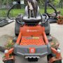 A small brindle and white dog stands confidently on the seat of a bright orange Husqvarna zero-turn lawn mower, surrounded by grass clippings and garden pots on a concrete pad in a fenced backyard.